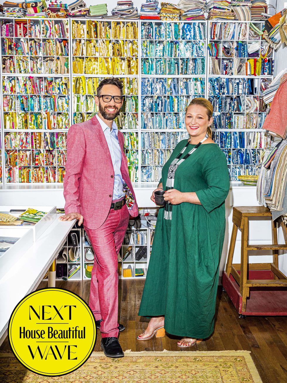 Matt and Heather French posing in front of wall of shelves filled with rainbow colored textiles with logo for Next Wave House Beatiful