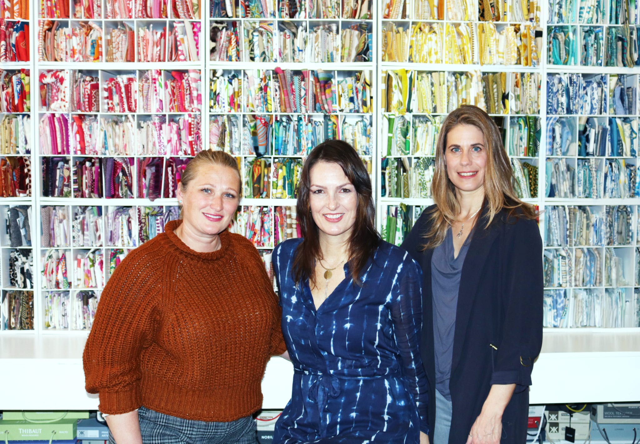 Heather French, Amy Flurry and Franziska Neumann posing for photo with rainbow colored fabric samples in shelves in background